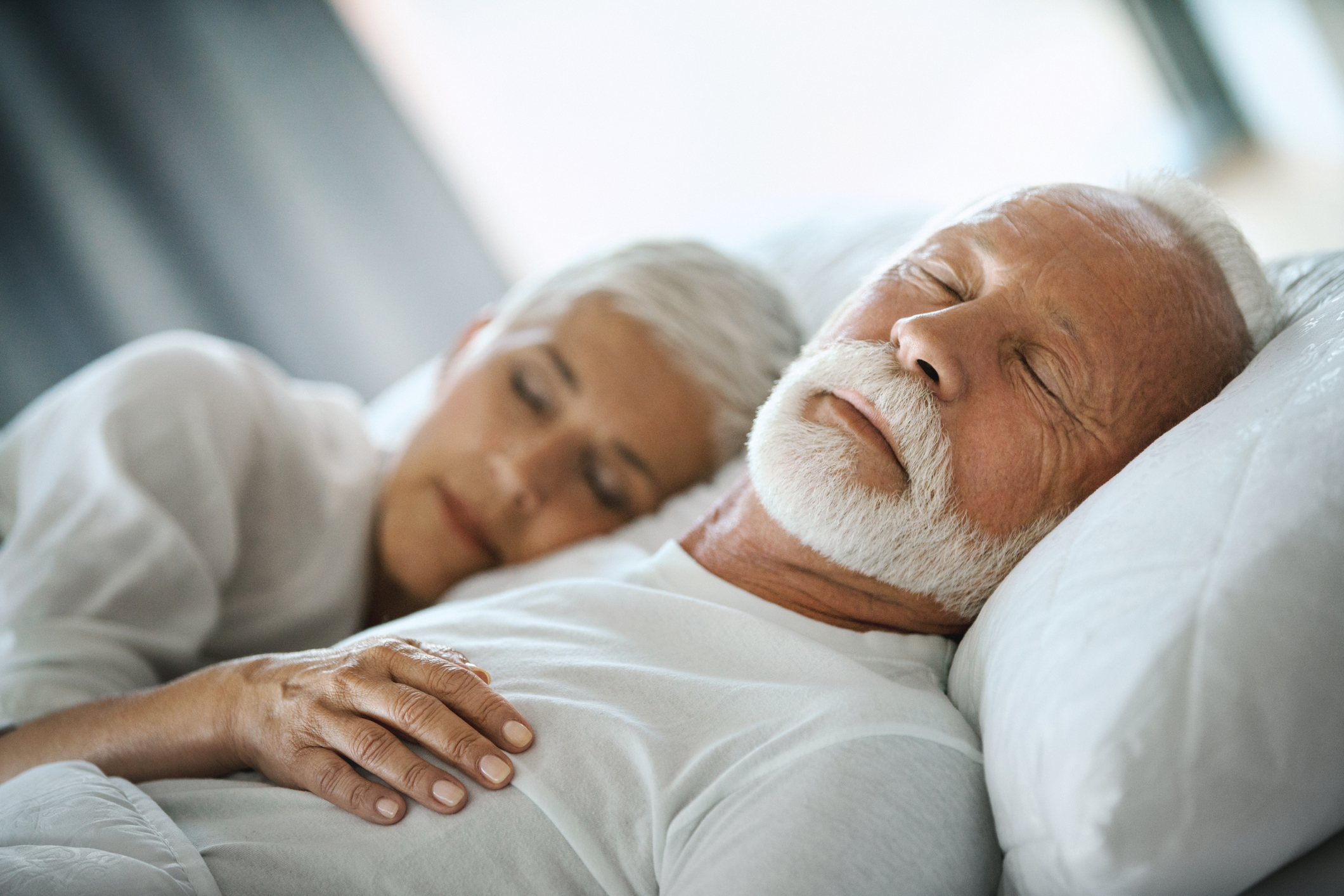 Closeup of mid 60's couple sleeping late in the morning in an already brightly lit bedroom.