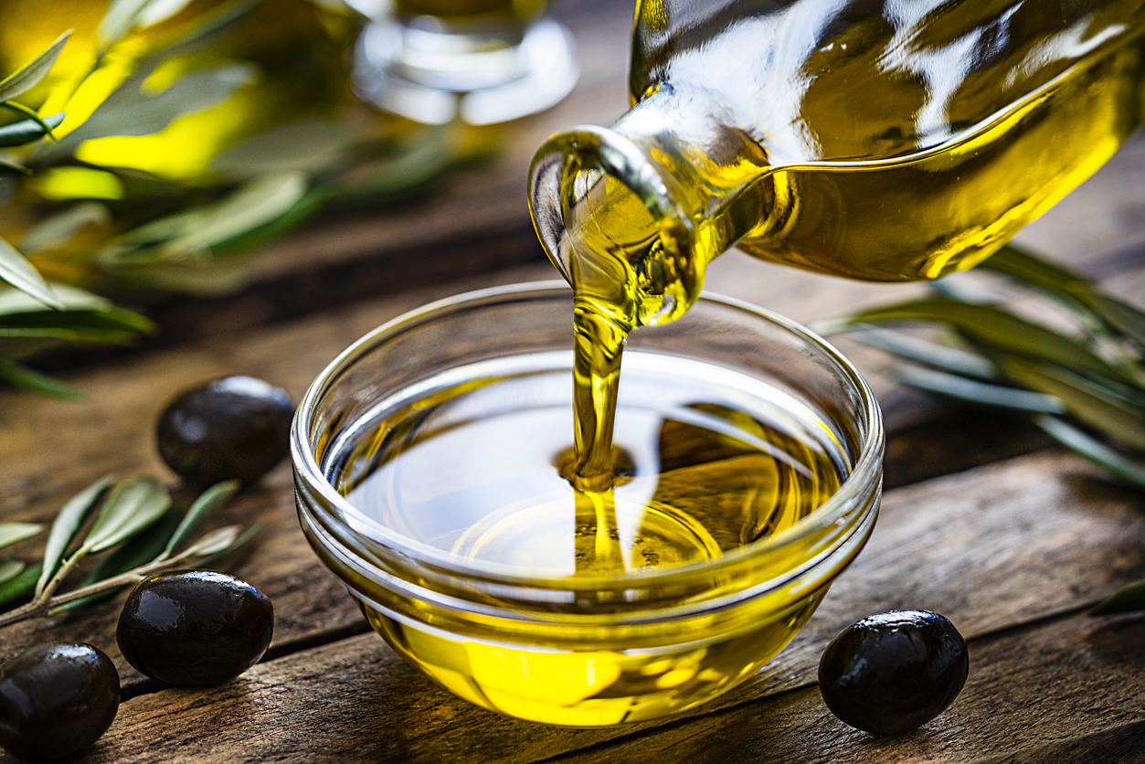 Pouring extra virgin olive oil from a bottle to a glass container. Olive branches comes from the left and right. Some olive oil bottles are out of focus at background. Black olives are on the table and complete the composition. The composition is on a rustic wooden kitchen table. Predominant colors are gold, green and brown. High resolution 42Mp studio digital capture taken with Sony A7rII and Sony FE 90mm f2.8 macro G OSS lens