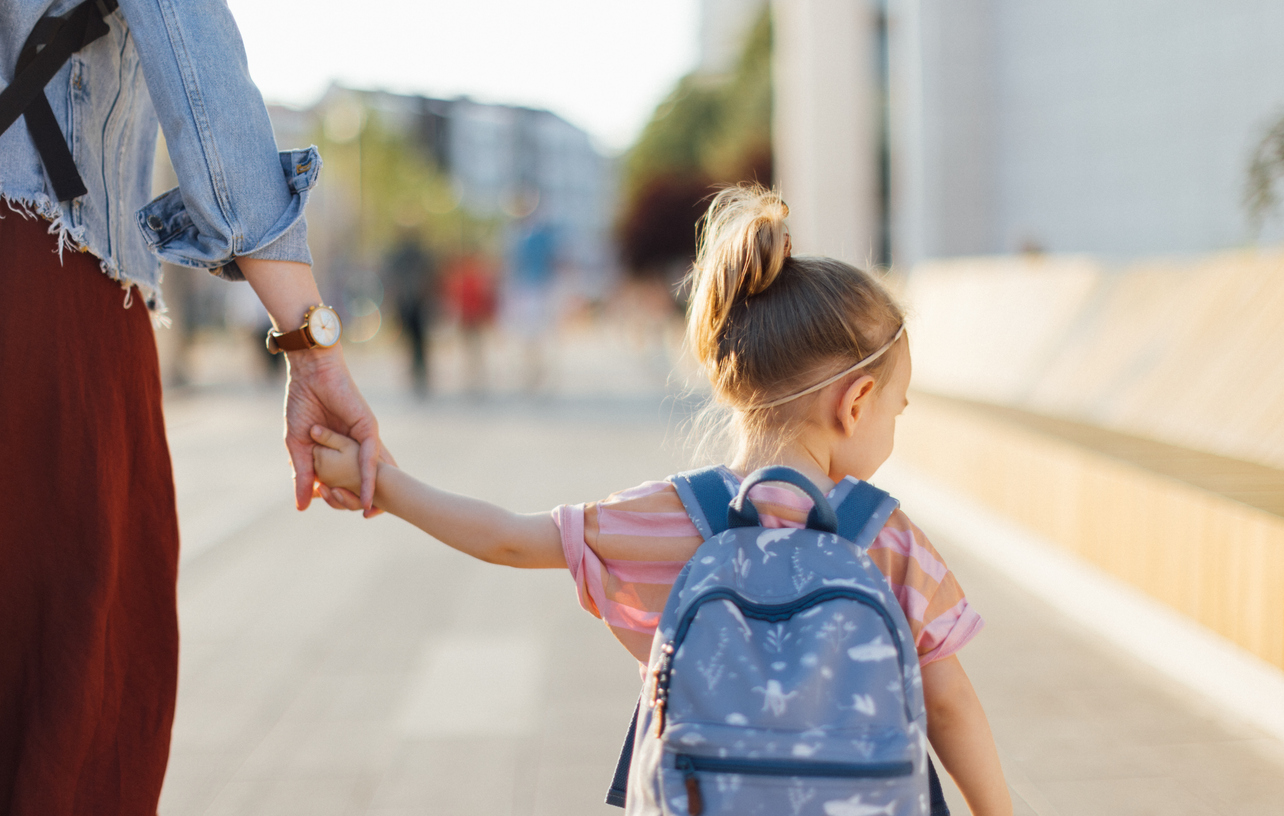 Back view of little girl with backpack holding a mom hand while walking to the school together.
