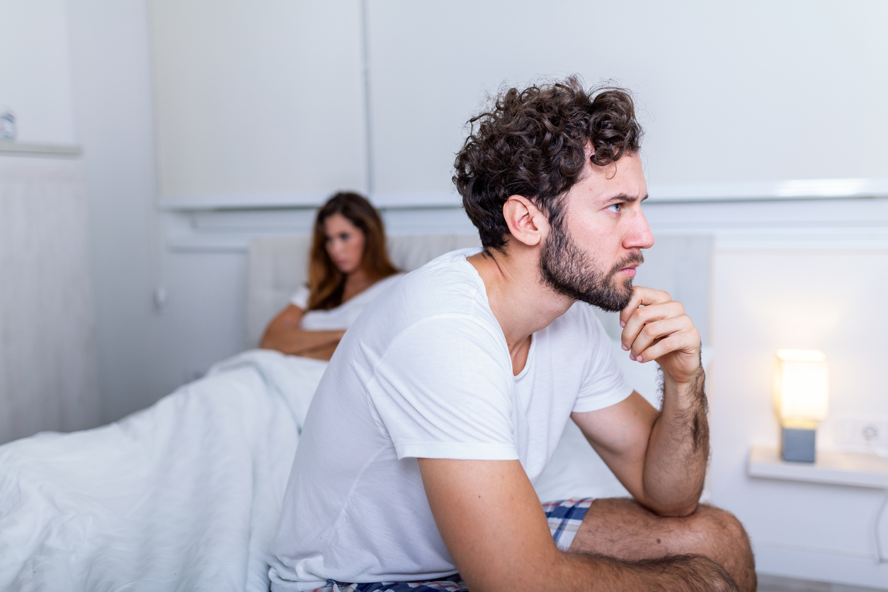 Sad thoughtful man after arguing with girlfriend.Relationship difficulties, conflict and family concept,unhappy couple having problems at bedroom. Sad guy sitting on bed,girlfriend in the background