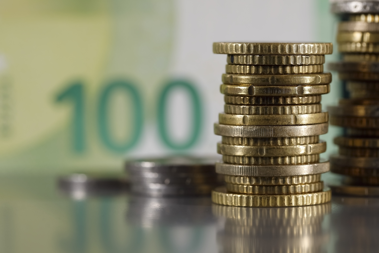 These are euro coins which have been stacked on a grey, flat surface and are visible against a blurred 100 euro banknote.