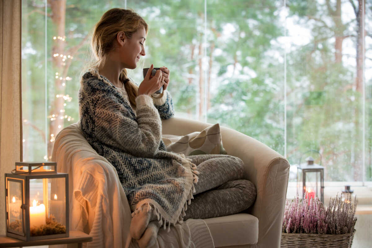 Young beautiful woman sitting home in the chair by the window with cup of hot coffee wearing knitted warm sweater. Cozy room decorated with lanterns and candles.