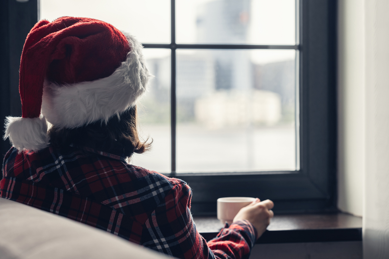 Back view of young woman in a red santa claus christmas hat sitting near window, having breakfast with cup of coffee. Concept