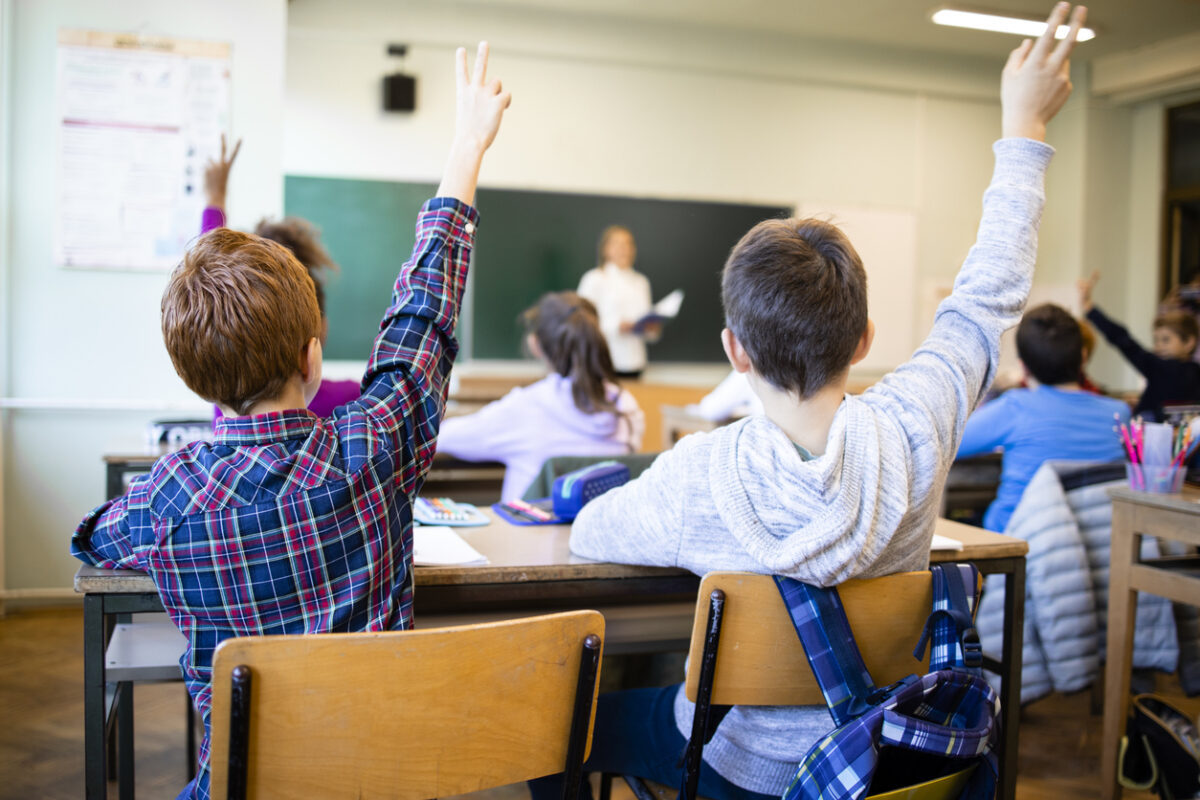 Schoolchildren at classroom with raised hands answering teacher's question.