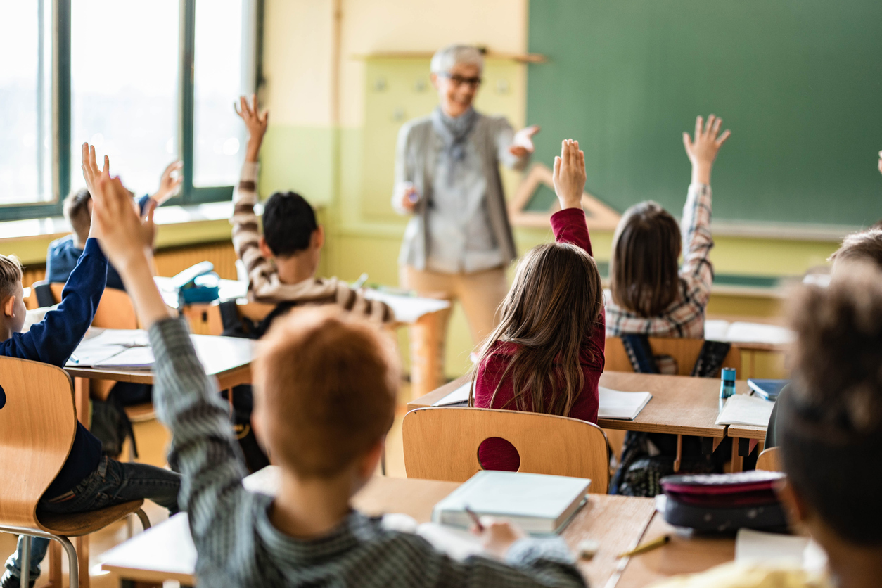Rear view of large group of elementary students raising hands to answer teacher's question on a class at school. Focus is on girl in the middle row in red t-shirt.