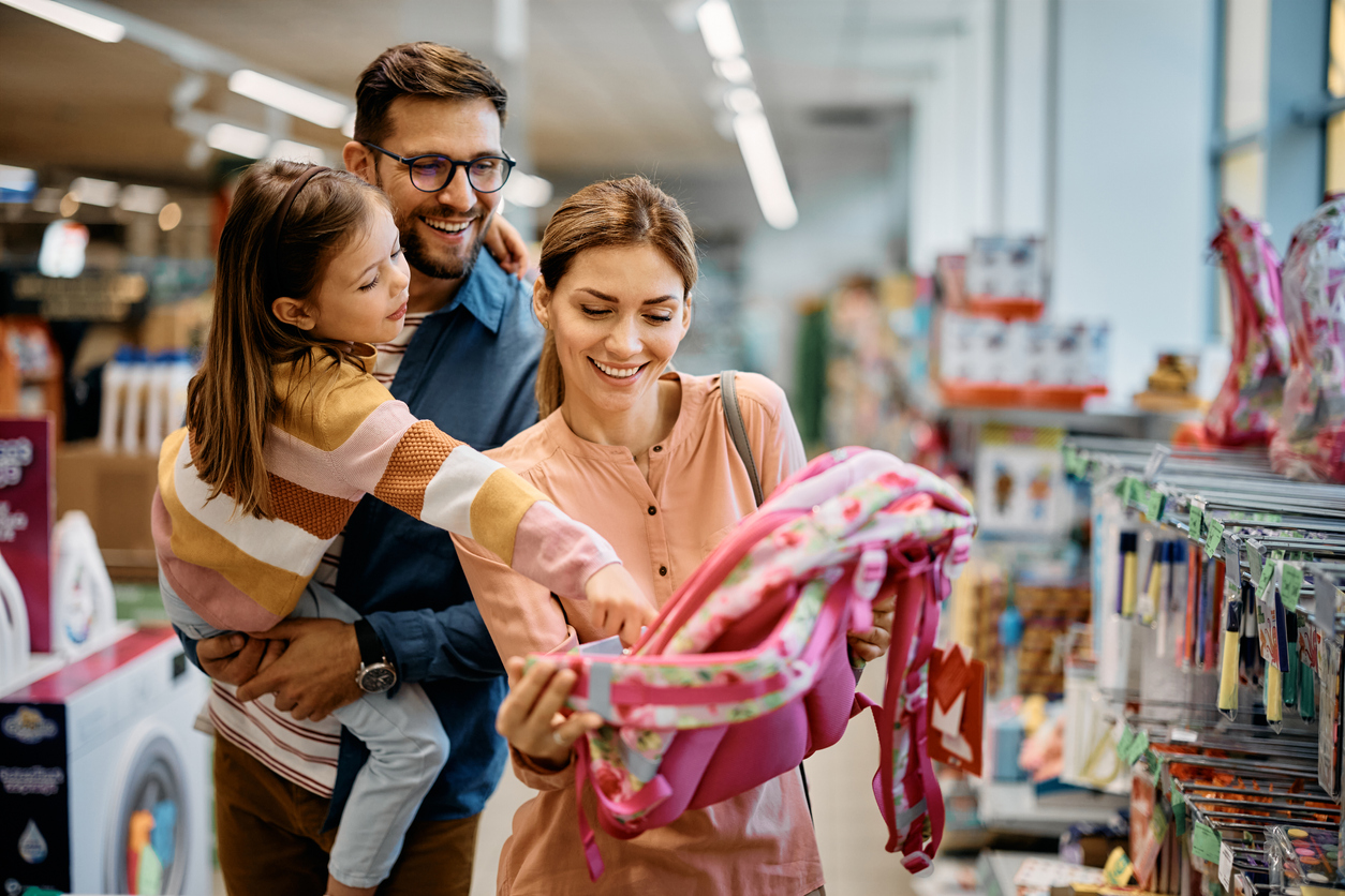 Little girl and her parents choosing backpack for school while shopping in the store together.