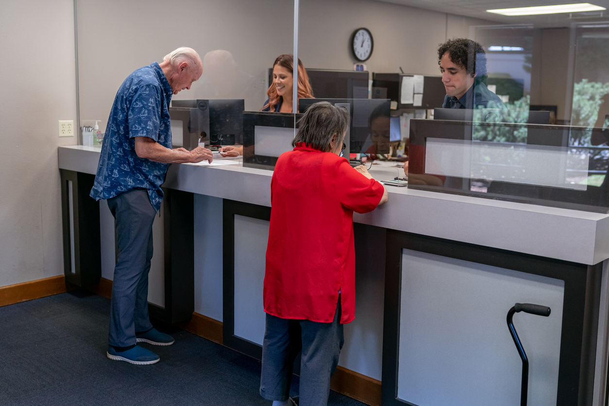 Two customer service representatives of Latin American descent assist happy senior adult customers while working at the reception desk of a medical office or bank.