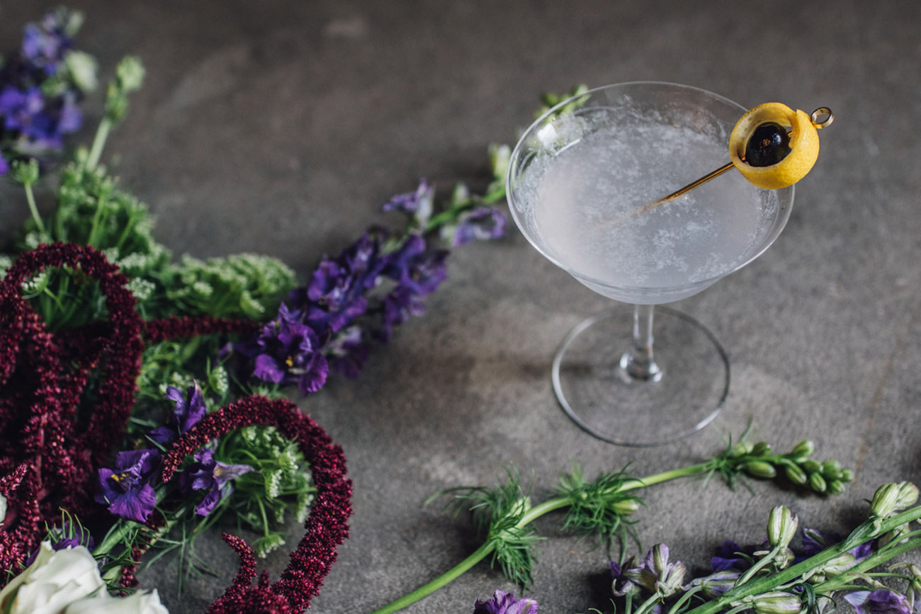purple gin Aviation cocktail with lemon and cherry garnish on concrete floor surrounded by greenery, floral, bouquet