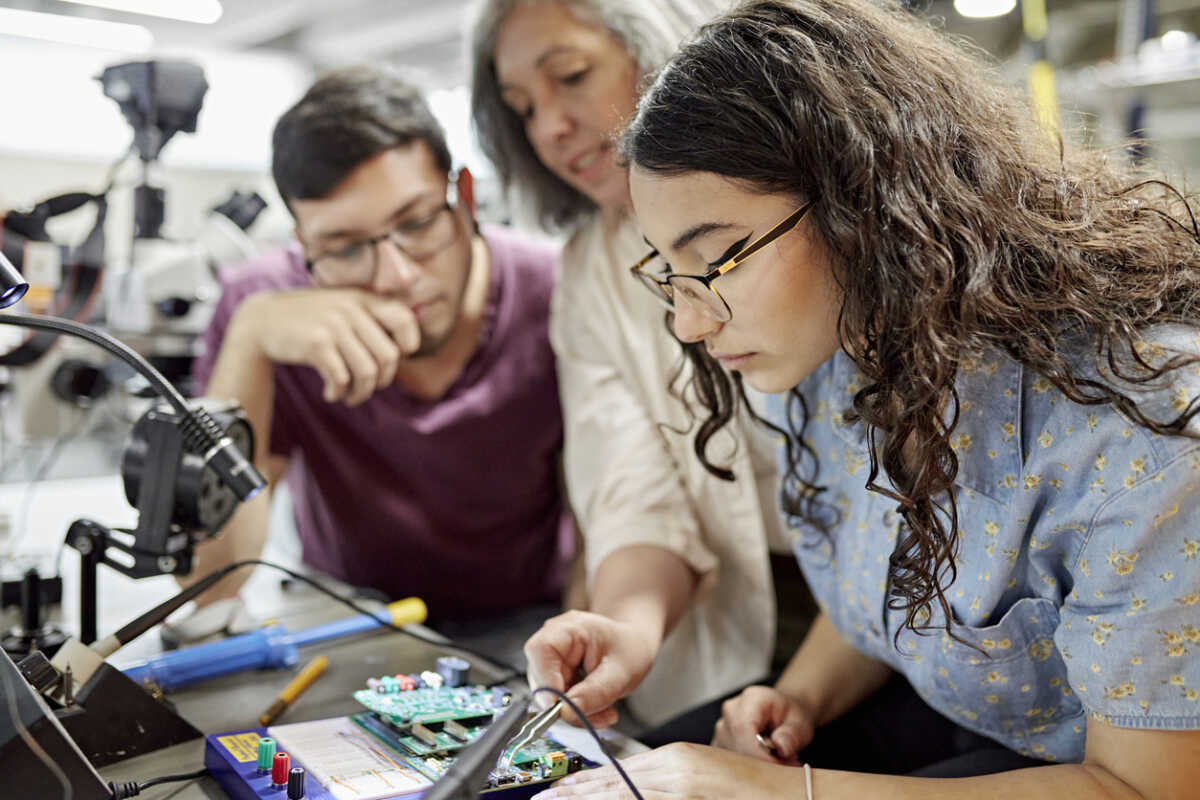 Close view of mature female educator standing between early 20s classmates and offering guidance as they work on project. Property release attached.