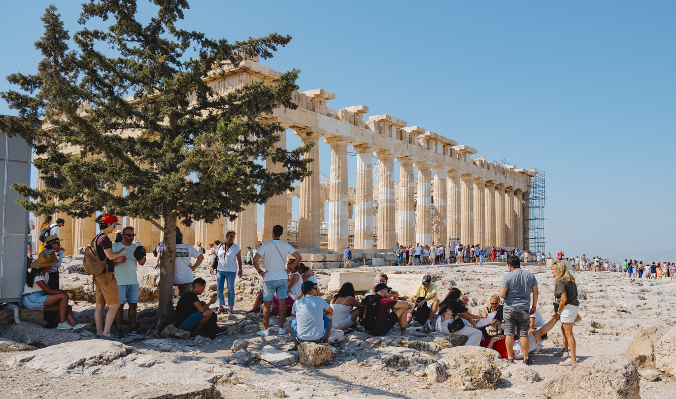 Athens, Greece - August 30, 2022: Visitors protecting themselves from the intense sun of a summer day in the shade of a tree, in front of the famous Parthenon, in the Acropolis of Athens, Greece