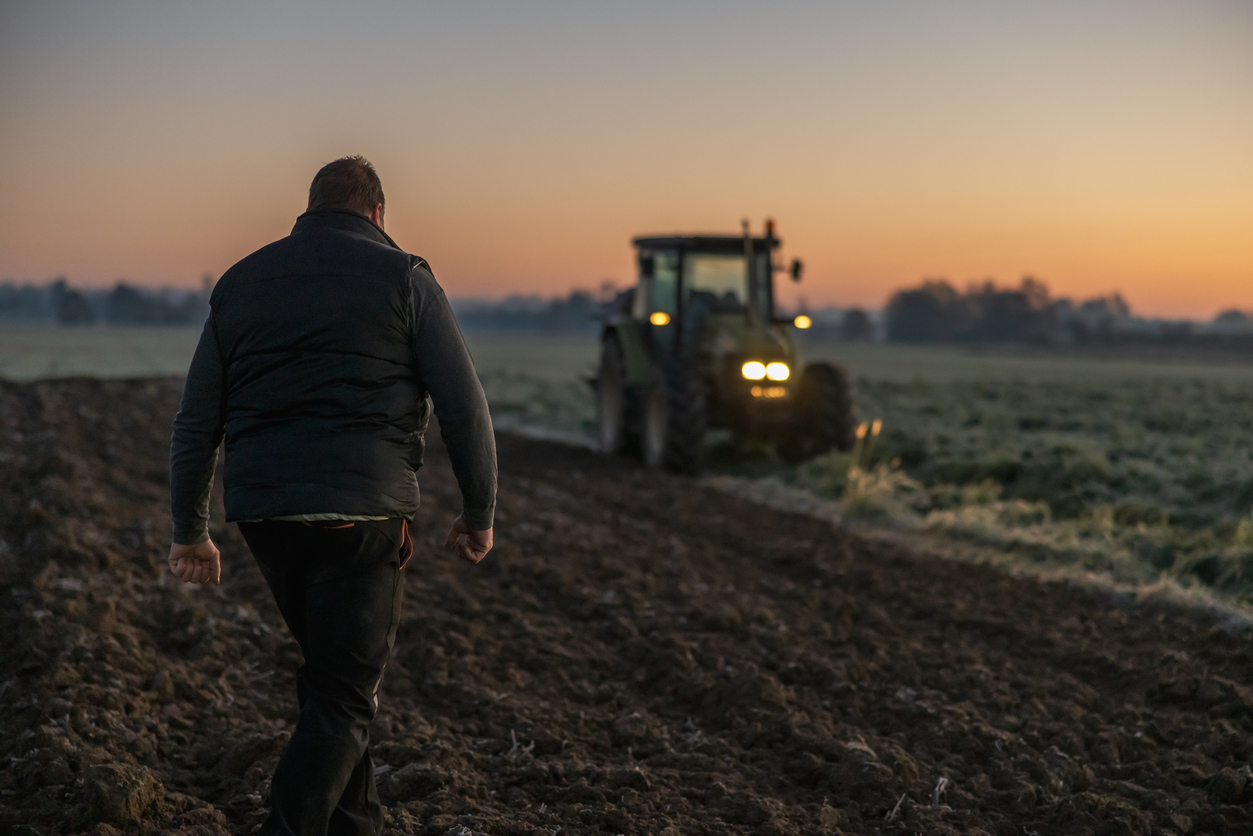 Man with short brown hair,wearing trousers and a black vest is walking on his agricultural field in the evening,rear view with green tractor in the background,lights on green tractor are turned on