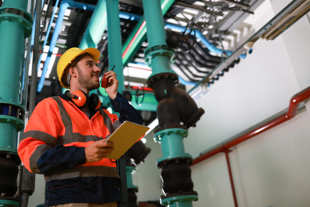 A young male engineer wearing an orange safety jacket is inspecting the piping system by walking around and checking it.