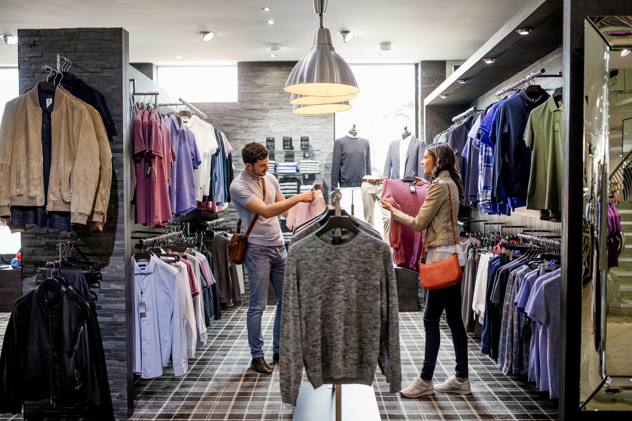 Young couple standing in clothes store with rails of shirts hanging around them. The man is holding a new pink shirt while the woman shows him another