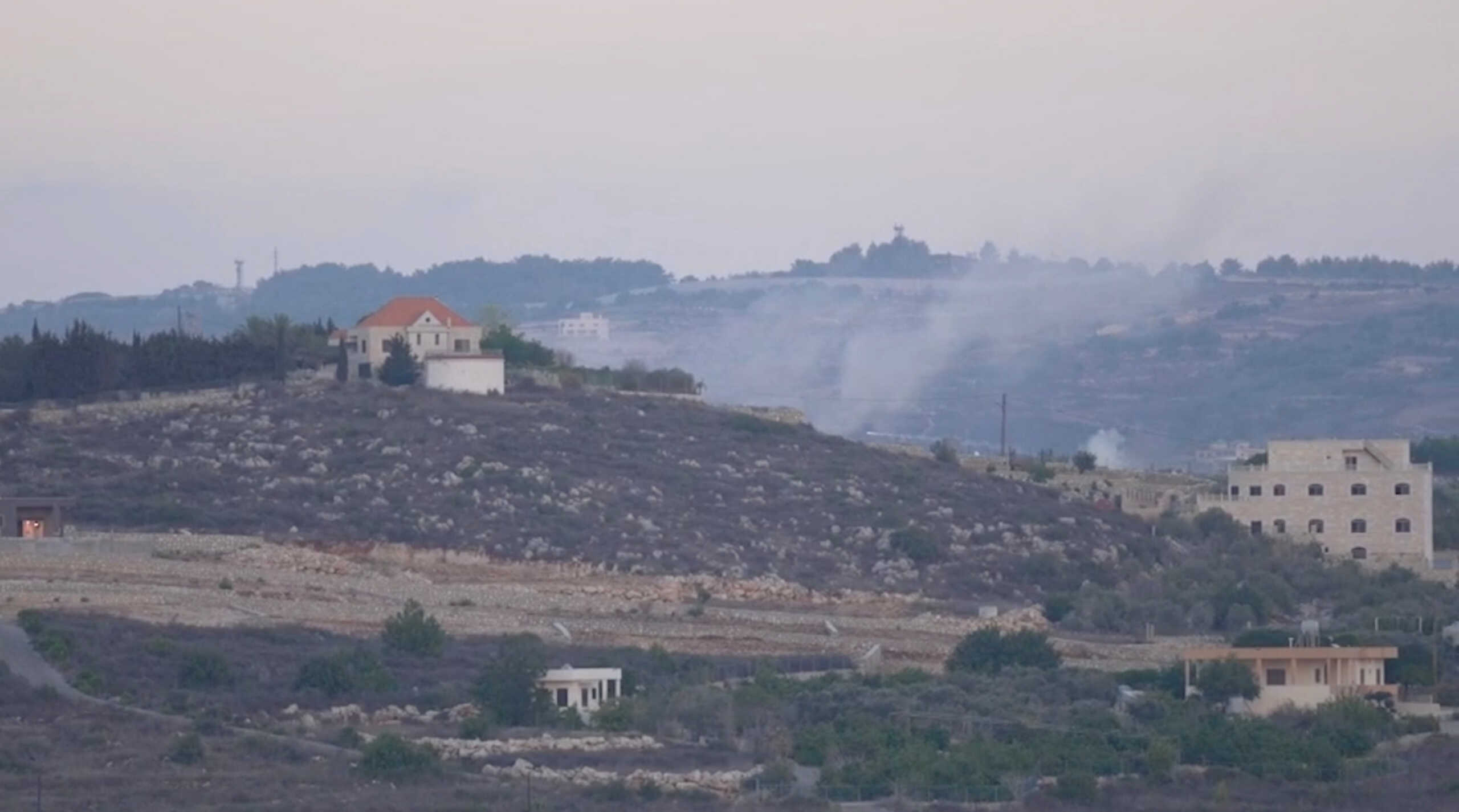 A screenshot from video footage provided to Reuters by Rai that was shot from the Lebanon side of the Israel-Lebanon border, October 13, 2023. The large plumes of smoke rising behind the hill to the right of the orange-roofed building indicate the location, close to the Lebanese village of Alma al-Chaab, that was hit by the first of two rounds fired by an Israeli tank, killing Reuters visuals journalist Issam Abdallah and wounding six other reporters. To match Special Report ISRAEL-LEBANON