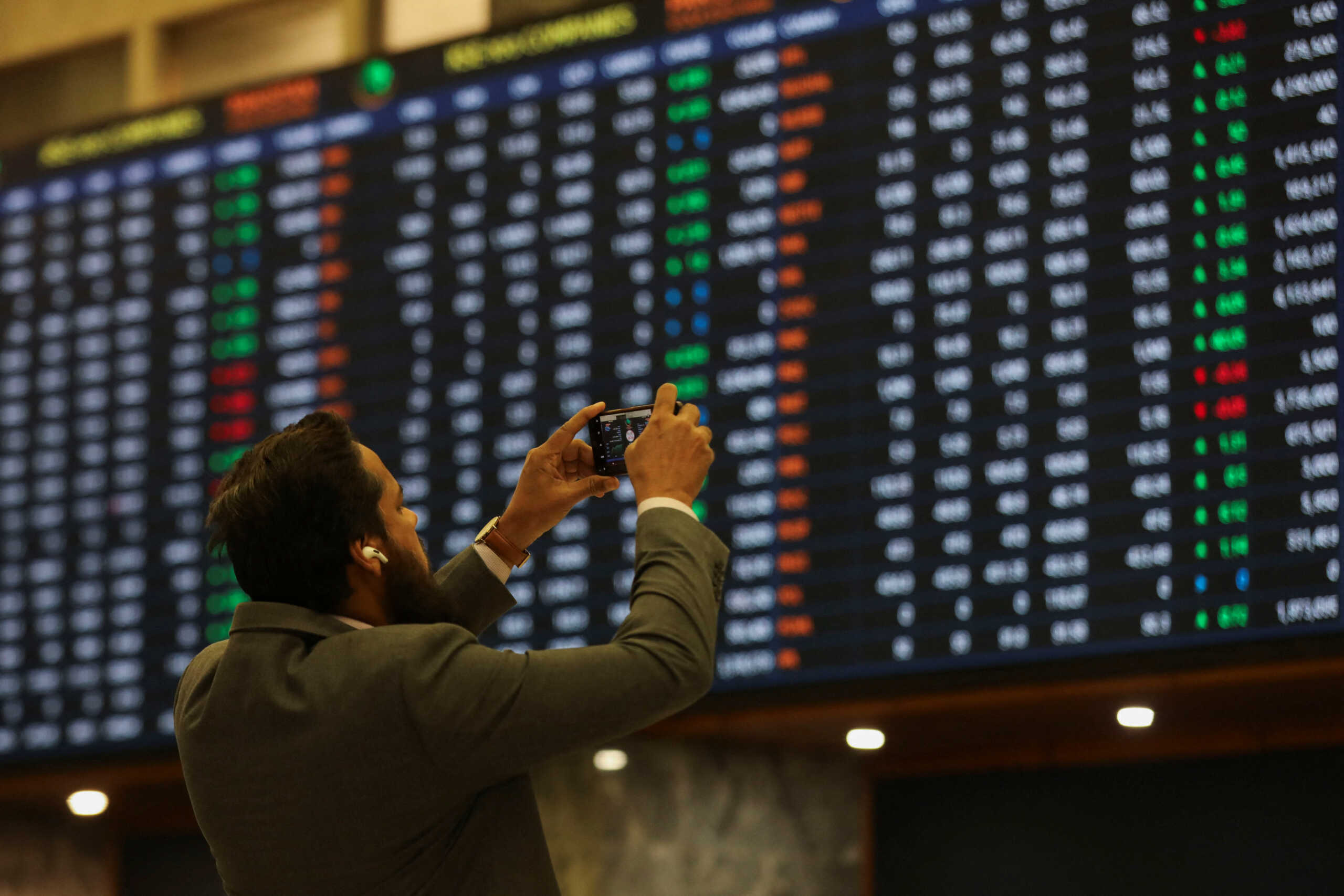 A man uses a mobile phone as he takes a photo of the electronic board displaying share prices during a trading session at the Pakistan Stock Exchange, in Karachi, Pakistan November 28, 2023. REUTERS