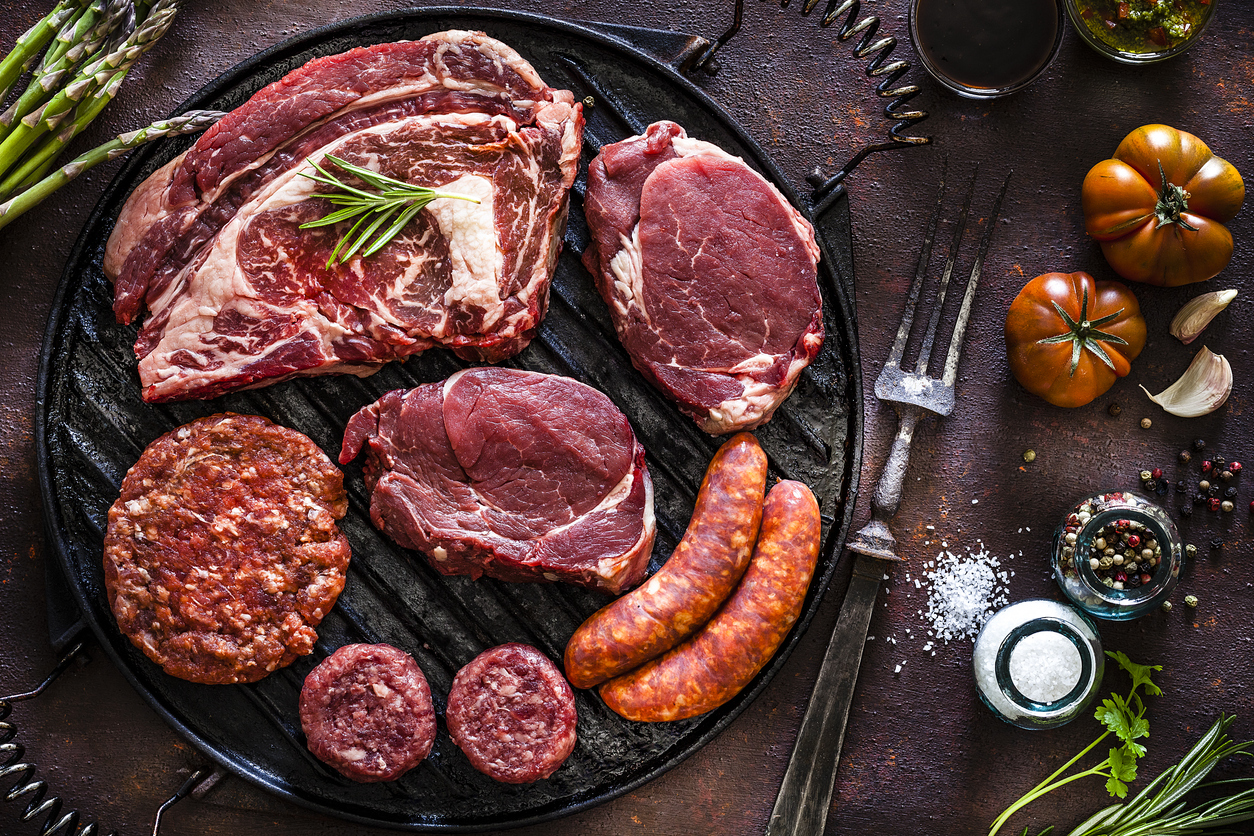 Various cuts of raw meat shot from above on rustic kitchen table. The cuts are on a cast iron grill and includes Angus steak, tenderloin, sausages and hamburger meat. The grill is surrounded by herbs, vegetables and spices for cooking meat like peppercorns, salt, garlic, tomatoes, asparagus and rosemary. A vintage fork is included in the composition. Predominant colors are red and brown. Low key DSRL studio photo taken with Canon EOS 5D Mk II and Canon EF 100mm f