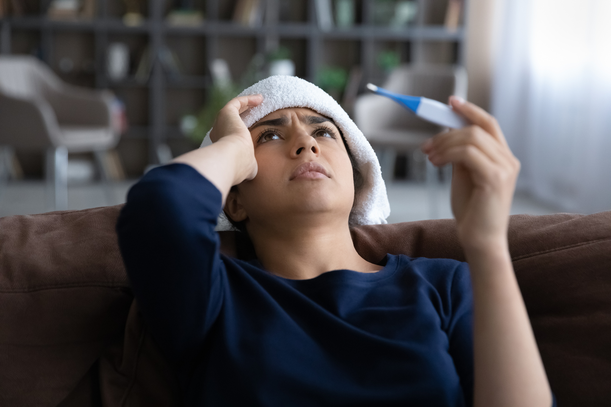 Close up sick Indian woman feeling unhealthy, holding thermometer, suffering from heat or fever, unhappy young female with towel on head leaning back on couch at home, cold or flu, health problem