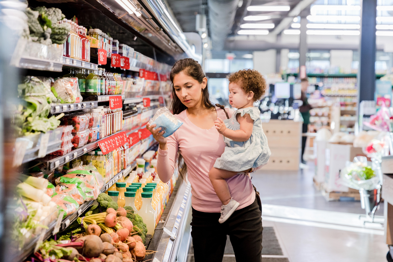 The young mother holds her daughter on her hip as she grocery shops for items she needs.