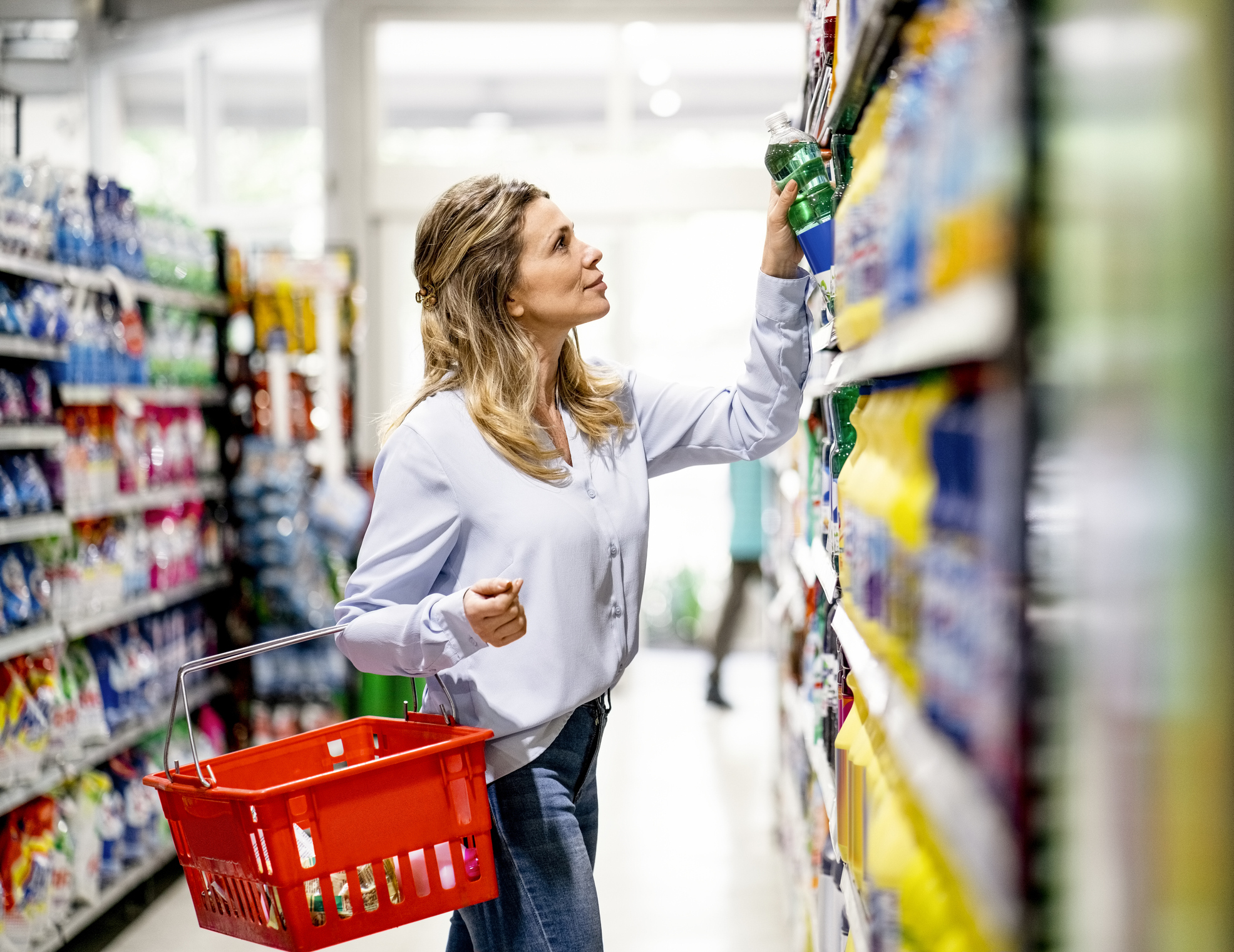 Woman customer buying groceries in supermarket . Woman with shopping basket picking up a product from display rack in retail store.