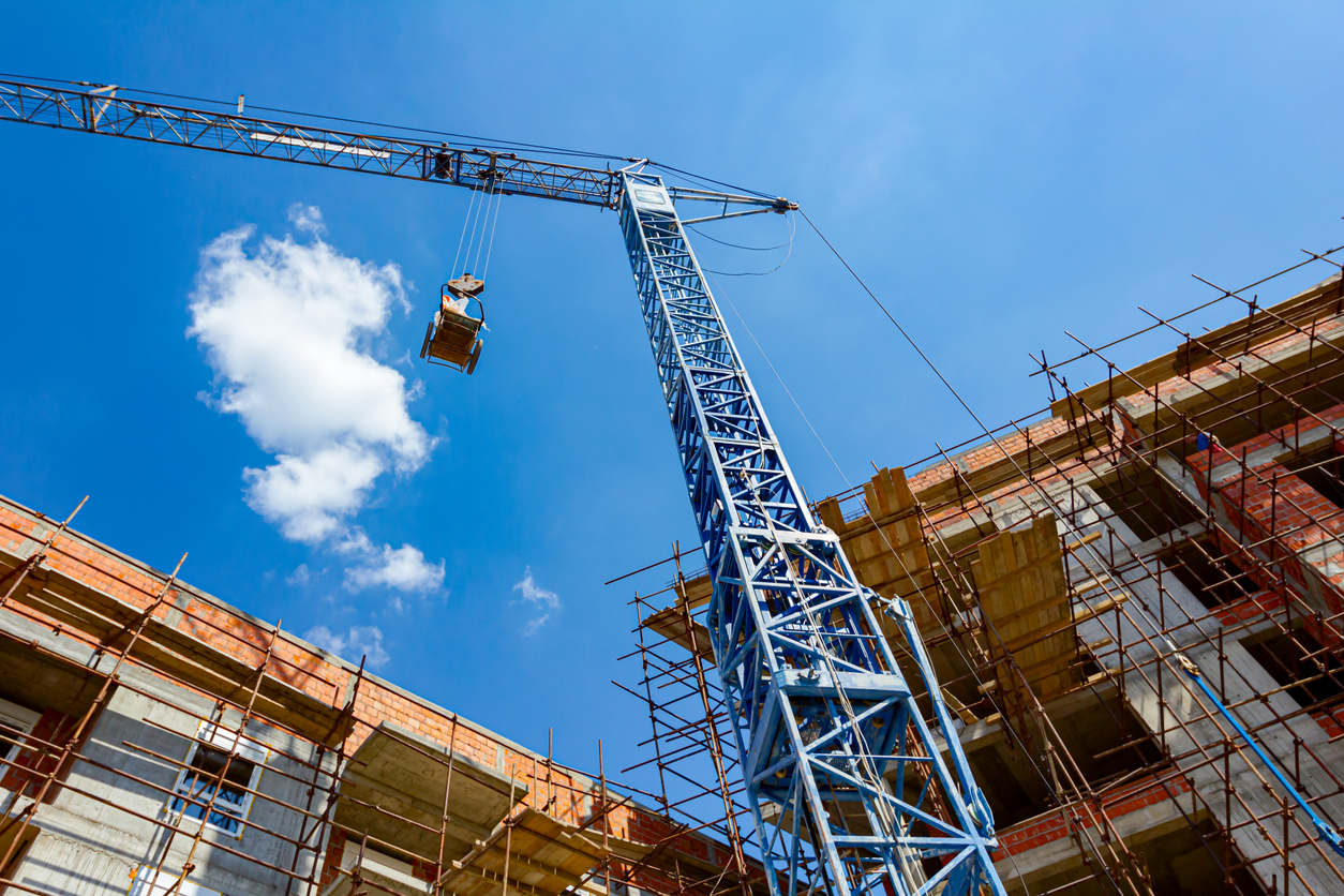 Shot from below of crane that puts down on ground wheelbarrow full of industrial waste, crumpled cellophane, in background is edifice under construction with scaffold and blue clear sky.