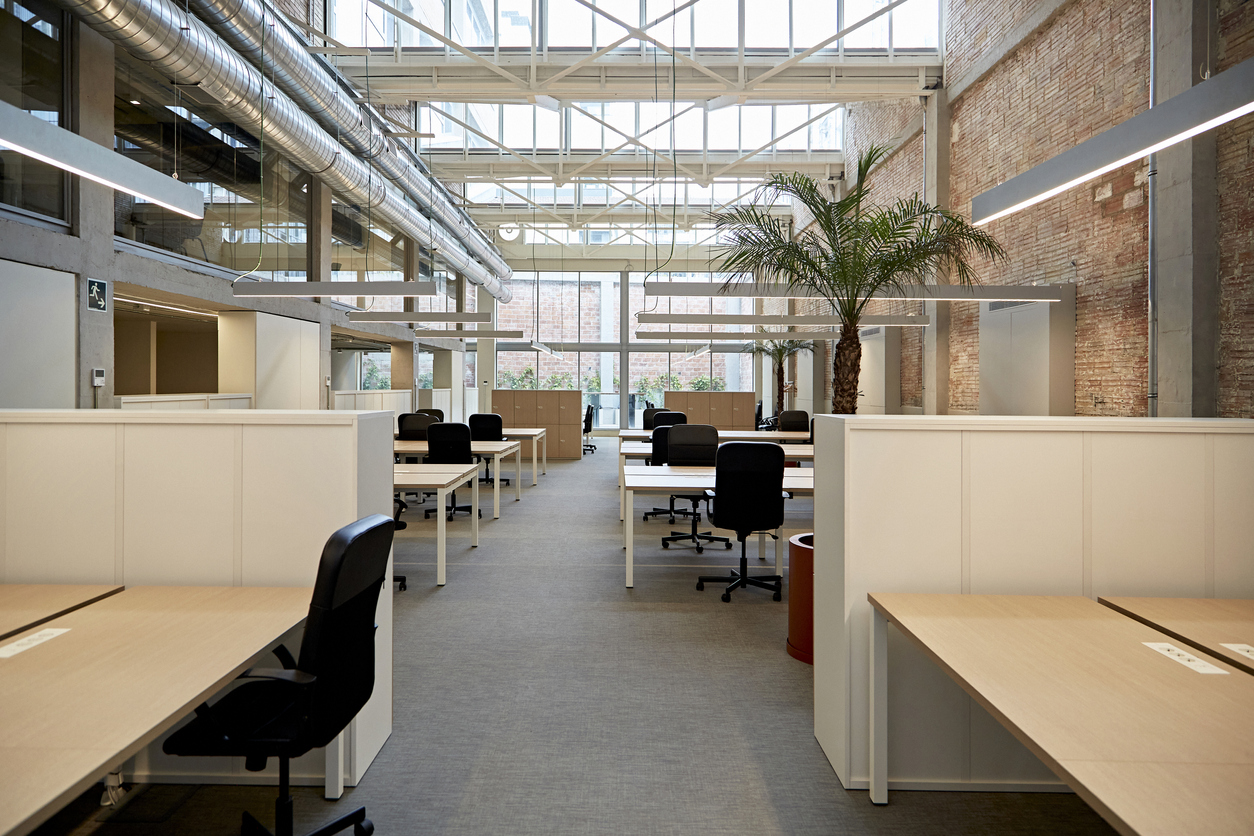 Wide angle view of unoccupied desks and chairs on ground floor of renovated building with exposed brick wall, high ceiling, and visible second floor windows.
