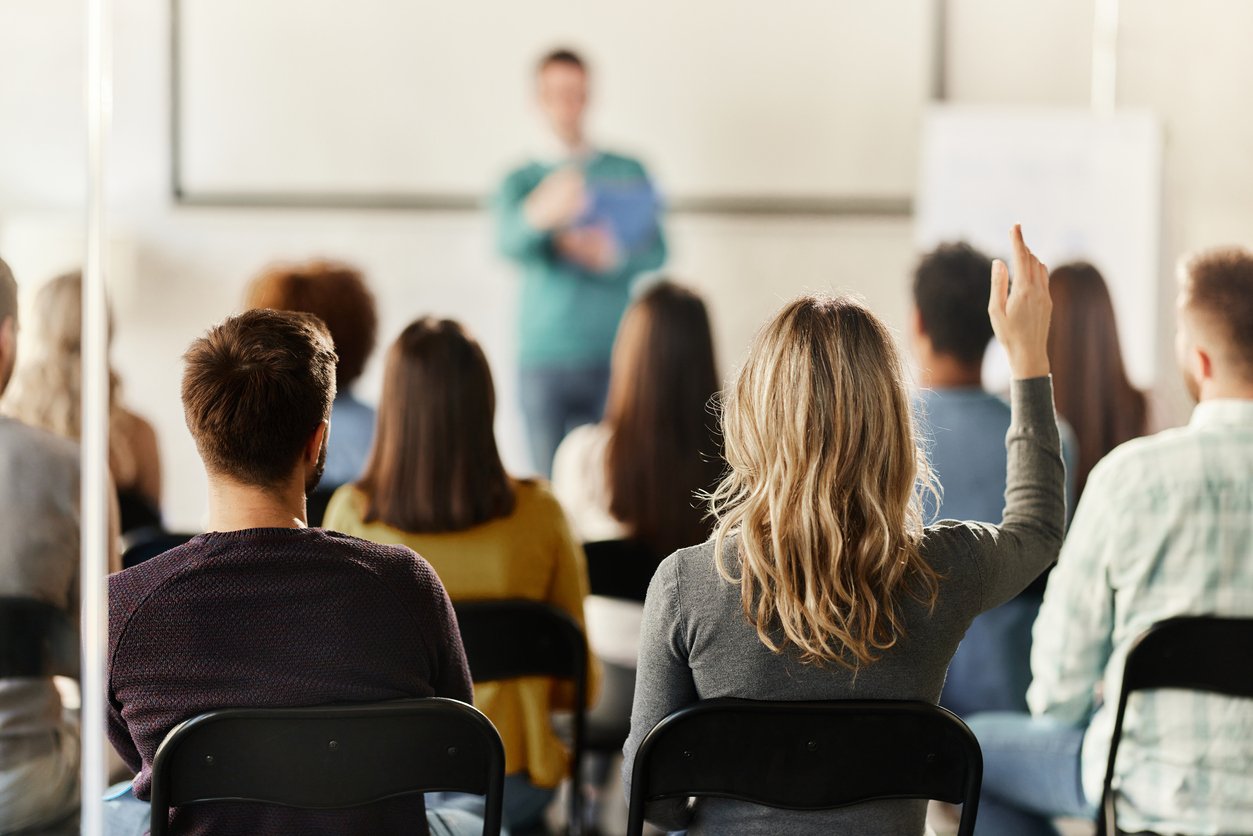 Back view of a businesswoman raising her hand to answer a question on a seminar in board room. The view is through glass.