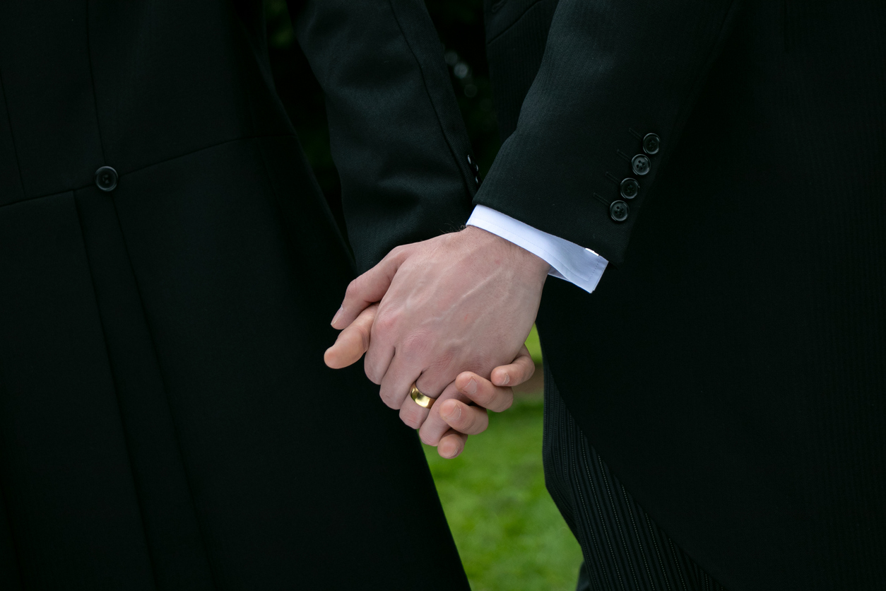 Gay wedding, married couple, men wearing mornings suits hold hands as they leave village church with wedding band on show