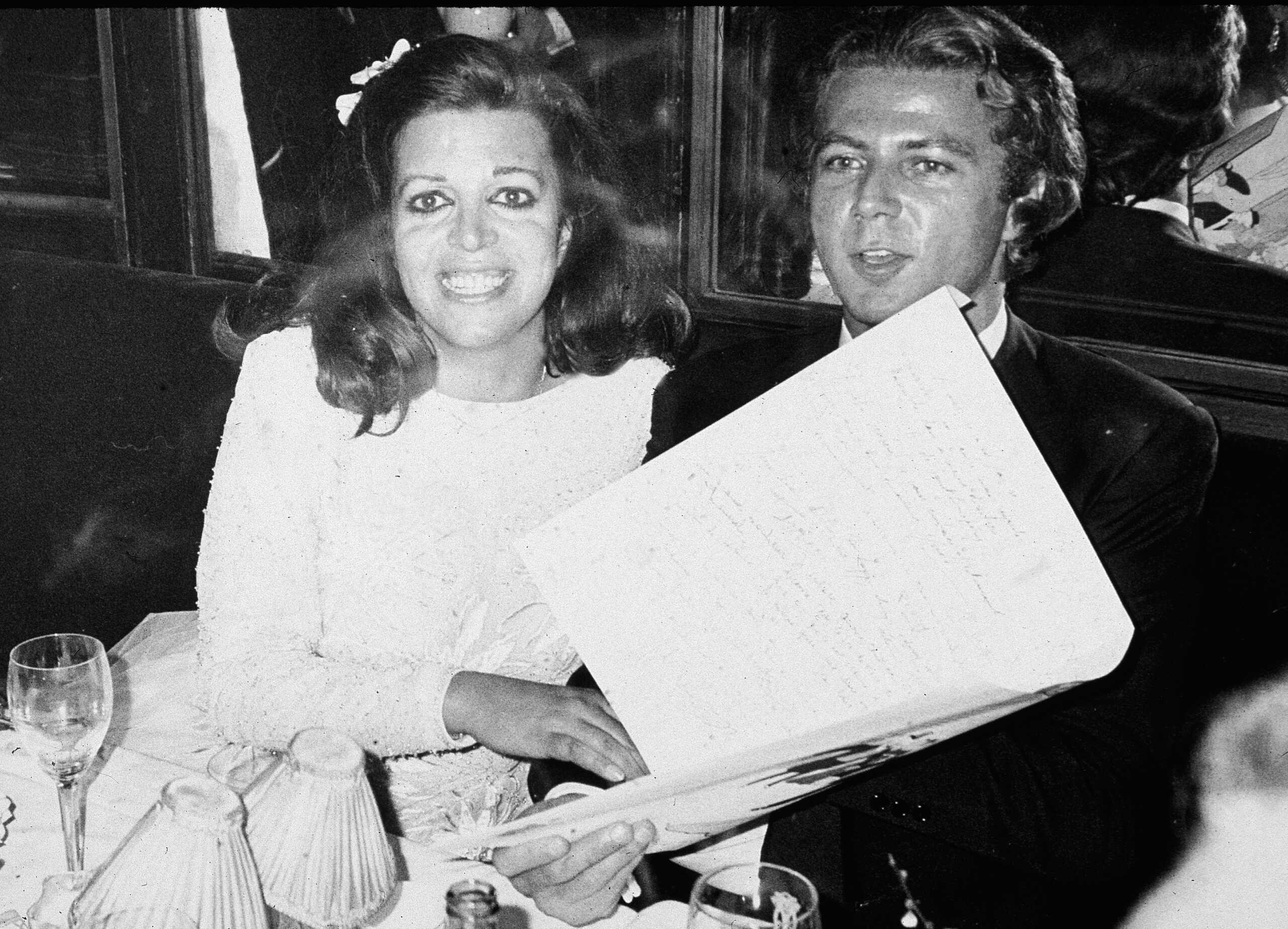 American-born shipping heiress Christina Onassis (1950 - 1988), the daughter of Greek shipping tycoon Aristotle Onassis, and her husband, French pharmeceutical heir Thierry Roussel, sit at a dining table and hold a large card at their wedding reception, Paris, France, March 1984. (Photo by Express Newspapers