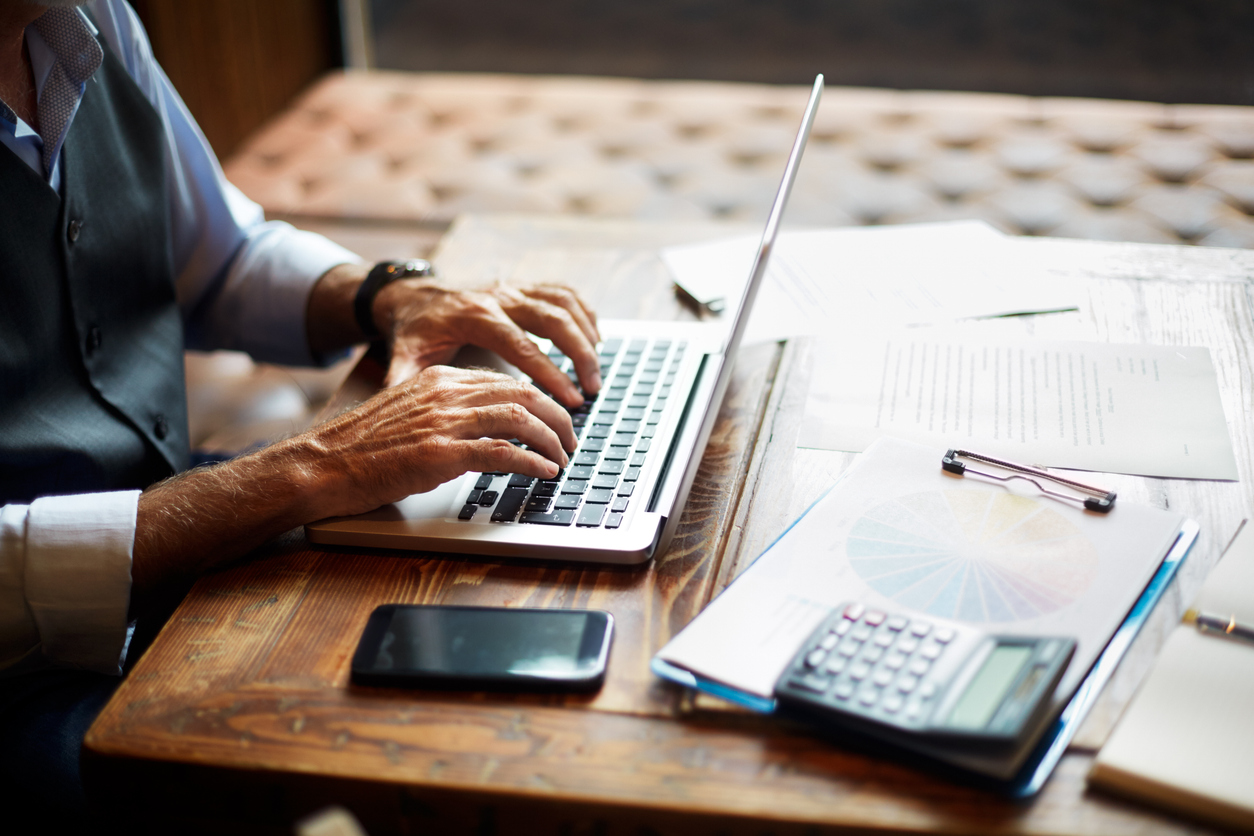Elderly unrecognizable entrepreneur using a laptop. Mobile phone, a calculator, a pie chart and some papers and notes are on the table.