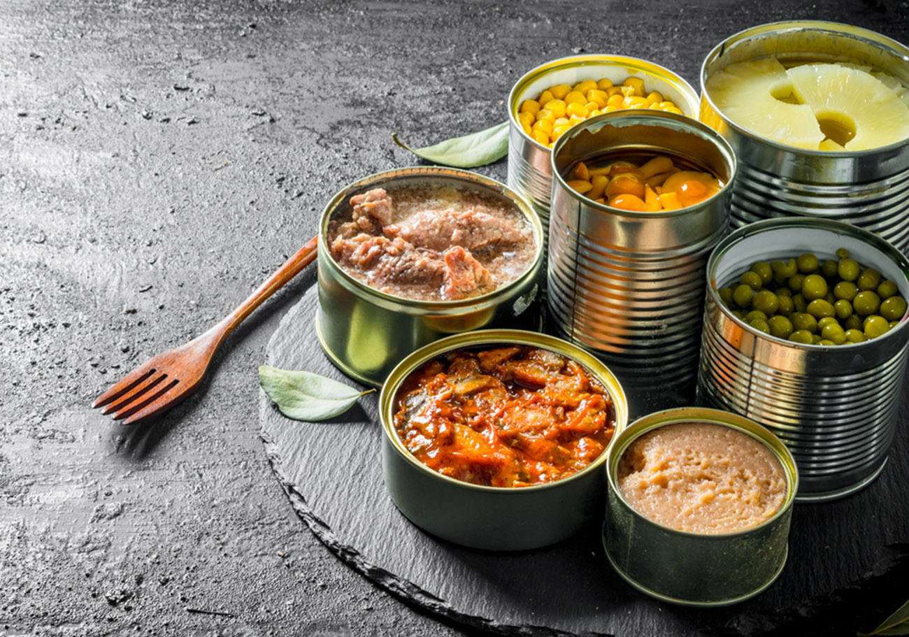 Various open tin cans of canned food on a stone Board with a fork. On black rustic background