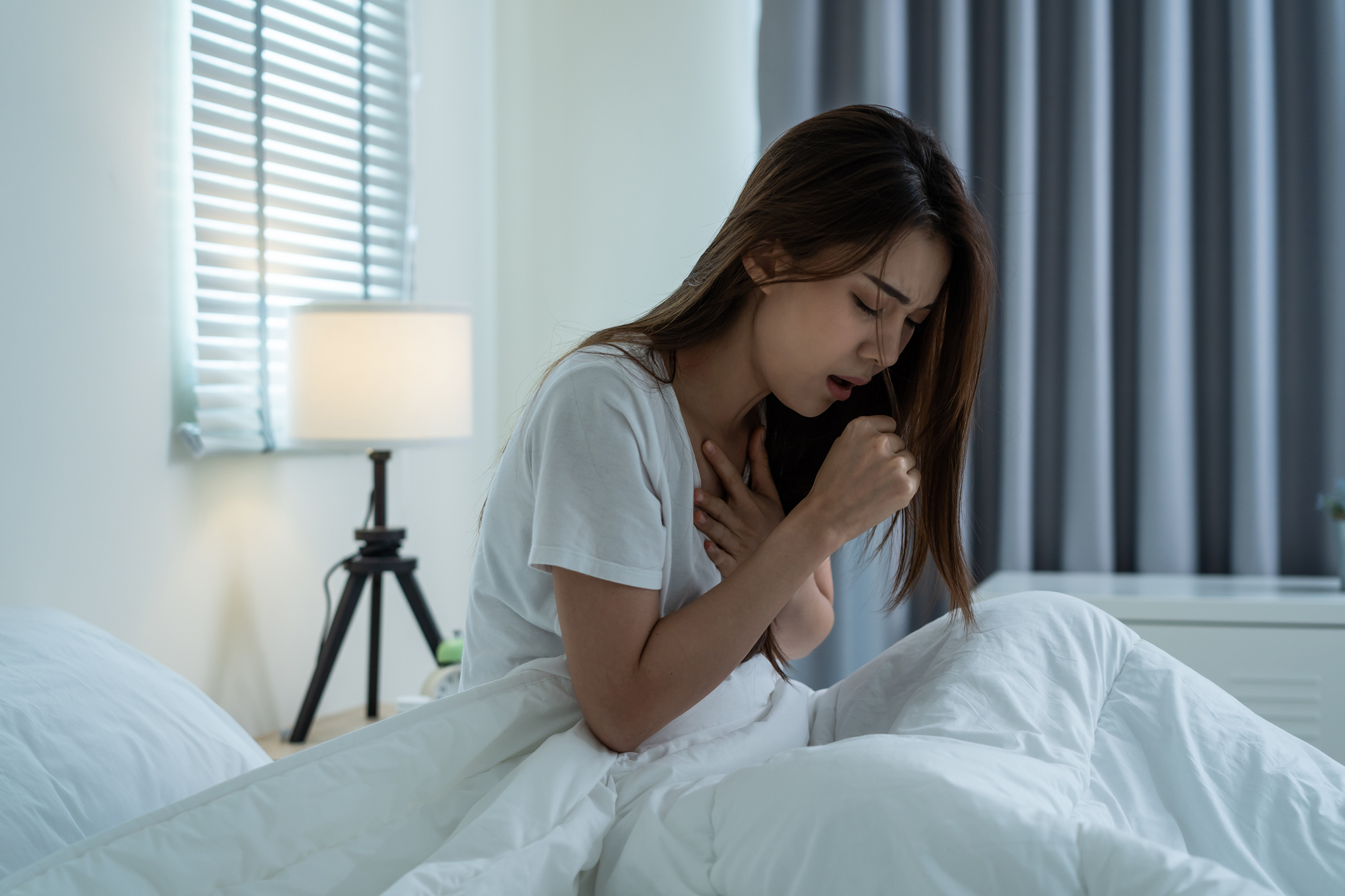 Asian beautiful girl in pajamas feel sick while sit on bed in bedroom. Attractive sleep young woman cover her mouth, feeling bad with fever and coughing after wake up on bed in the morning at home.