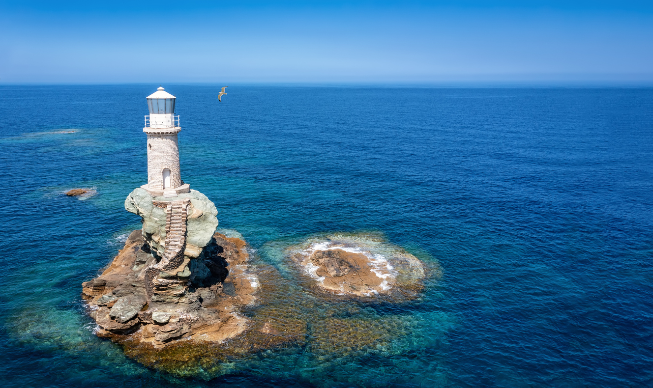 The Tourlitis lighthouse on a steep rock in calm, blue sea with a seagull, Andros island, Cyclades, Greece
