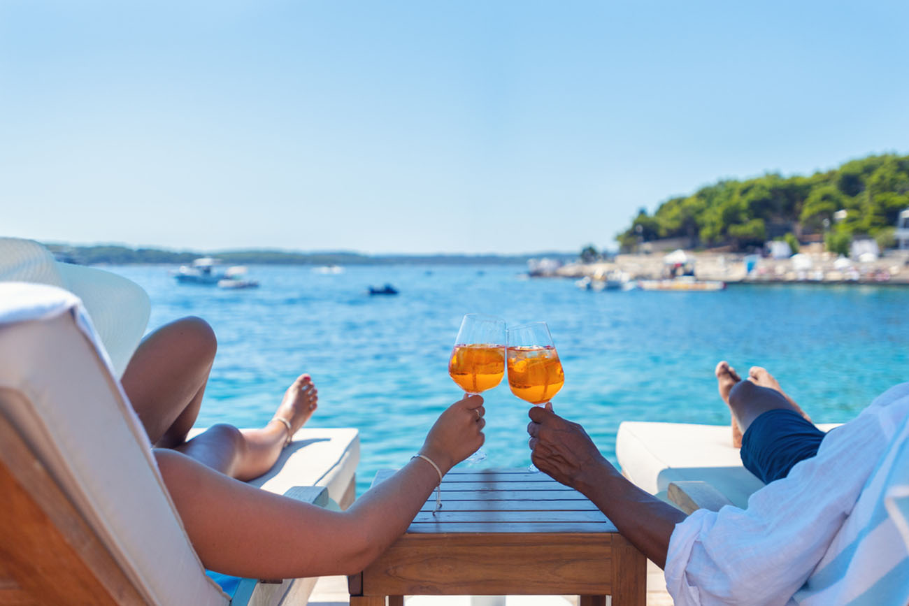 Couple relaxing and toasting with a Spritz cocktail on a beach deck over the ocean. Turquoise ocean is crystal clear.  horizon in the distance.