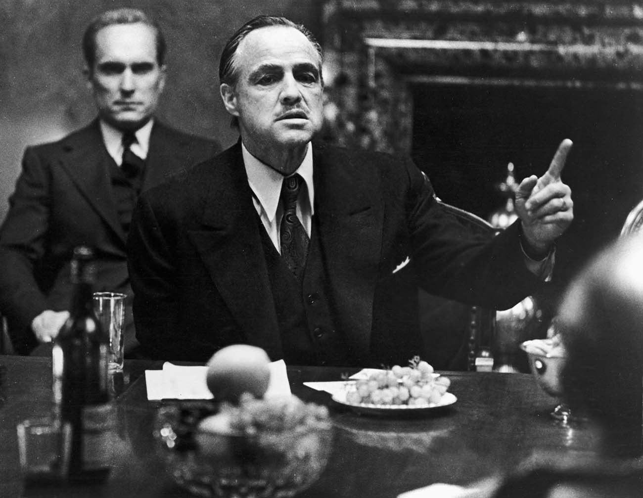 1972:  American actor Marlon Brando gestures at a table while American actor Robert Duvall sits behind him in a still from the film, 'The Godfather,' directed by Francis Ford Coppola and based on the novel by Mario Puzo.  (Photo by Paramount Pictures