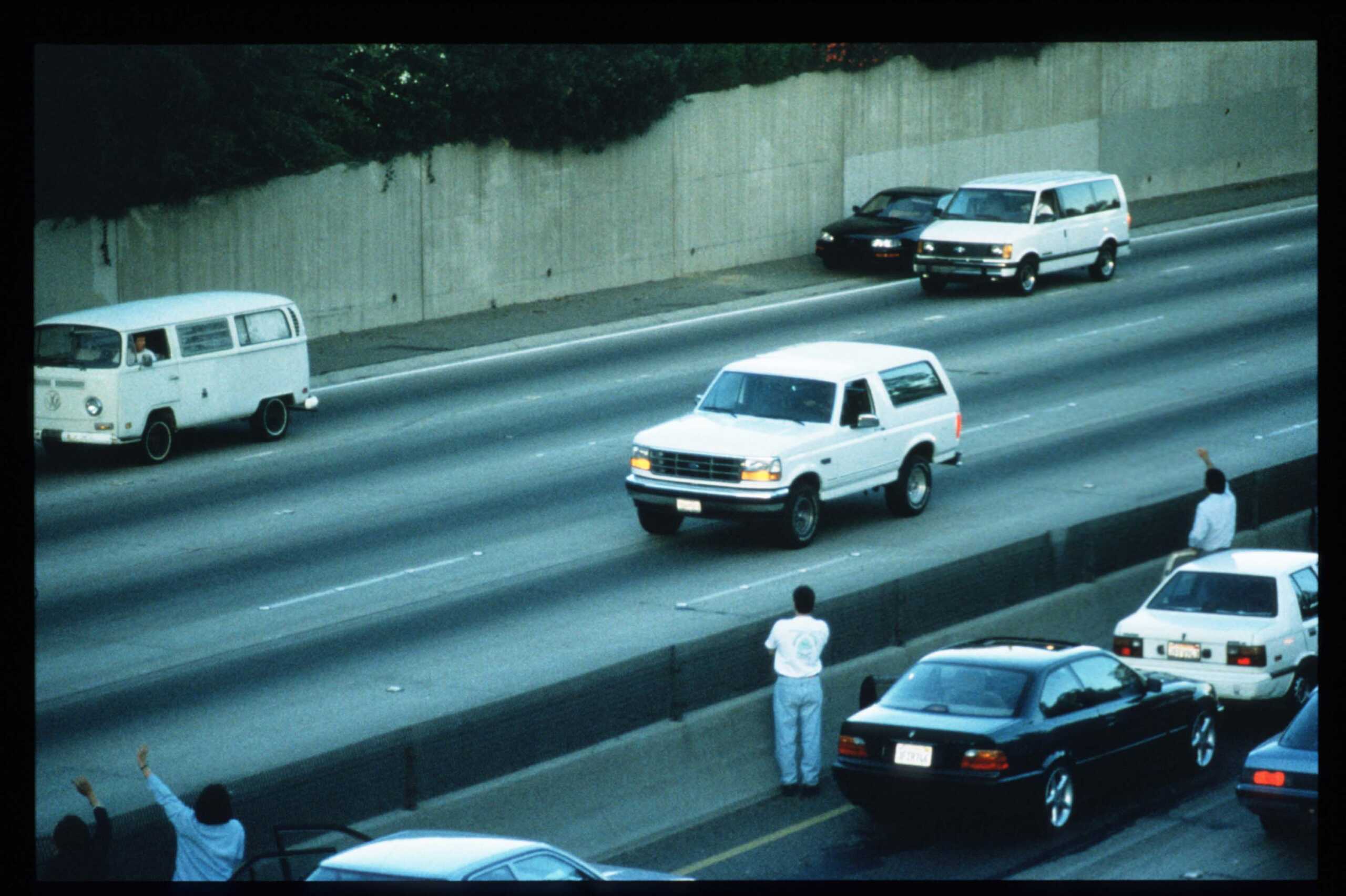 LOS ANGELES - JUNE 17:  Motorists wave as police cars pursue the Ford Bronco (white, R) driven by Al Cowlings, carrying fugitive murder suspect O.J. Simpson, on a 90-minute slow-speed car chase June 17, 1994 on the 405 freeway in Los Angeles, California. Simpson's friend Cowlings eventually drove Simpson home, with Simpson ducked under the back passenger seat, to Brentwood where he surrendered after a stand-off with police. (Photo by Jean-Marc Giboux