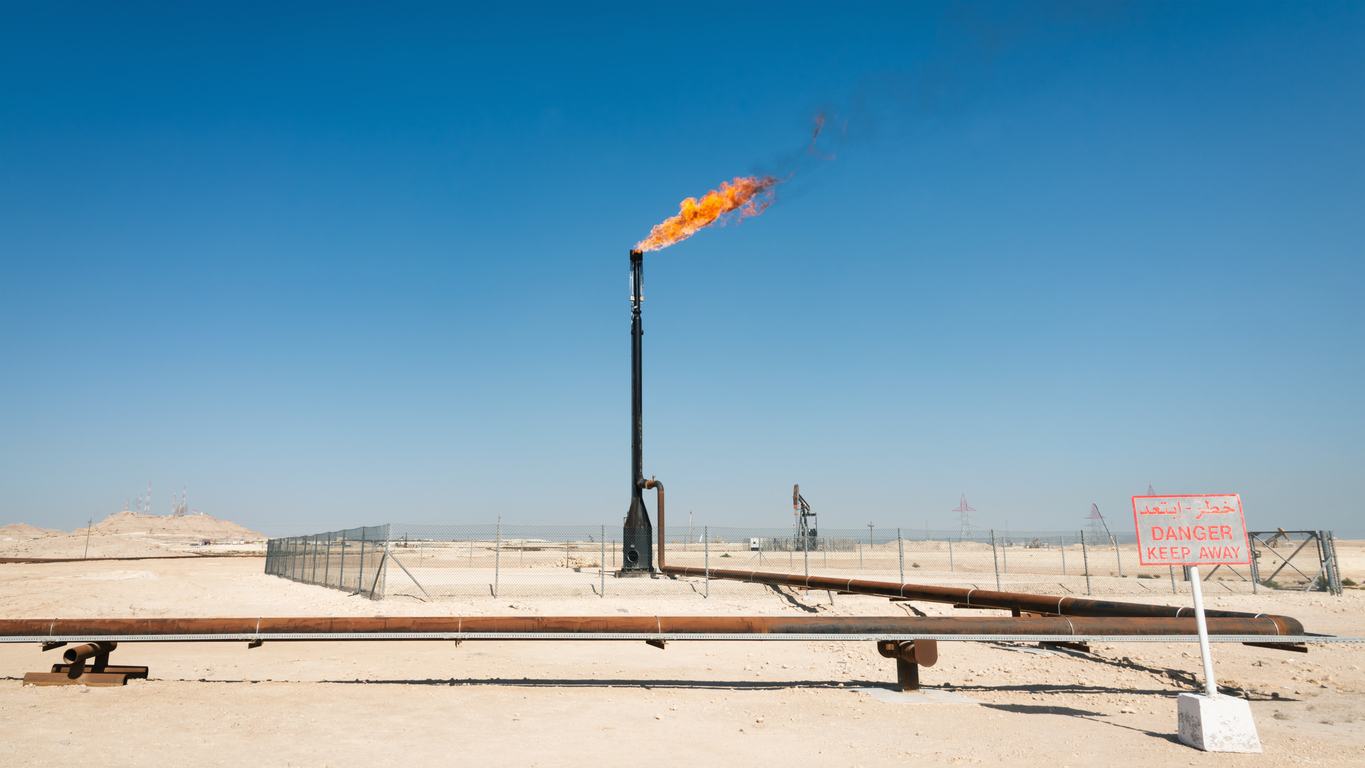Burning Flare Stack in Bahrain Middle Eastern Desert under blue summer sky. Natural Gas - Oil Industry Pipeline in the background. Danger Warning Sign on the Panorama left. Bahrain, Middle East, Persian Gulf Countries.
