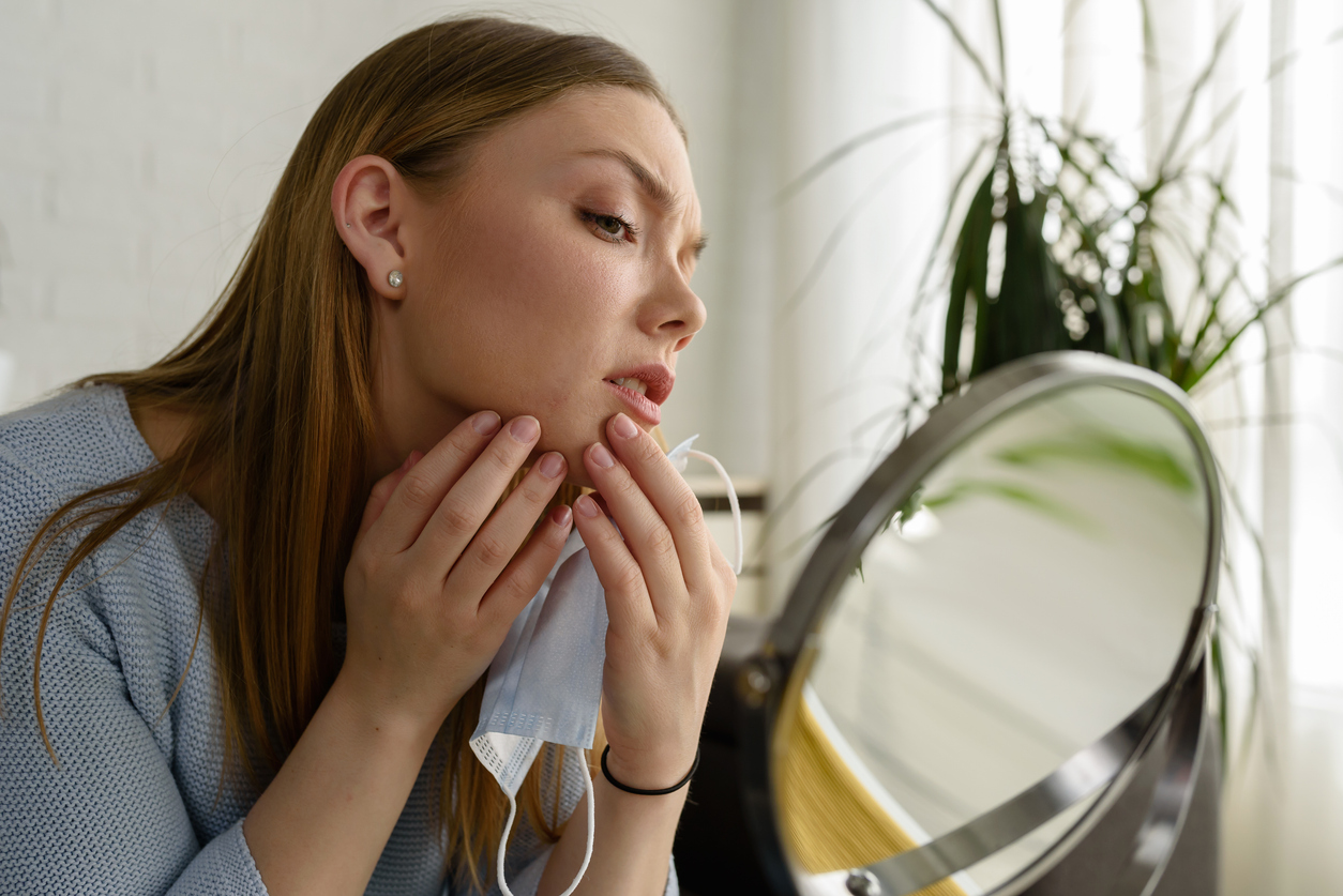 Young beautiful woman looking herself in the mirror at home. Worried about acne caused by wearing a mask. Maskne. Problems with acne.