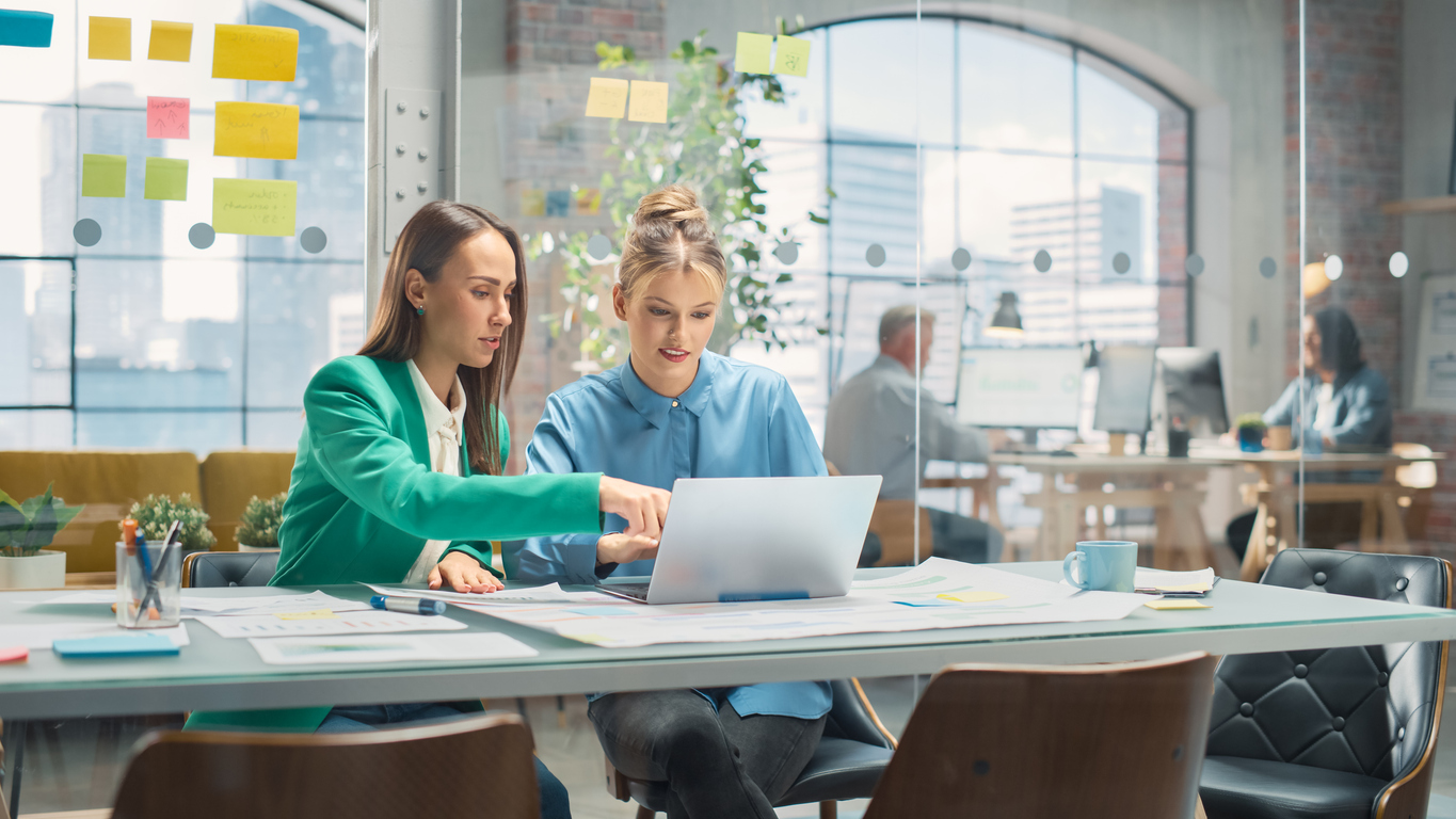 Portrait of Two Creative Colleagues Collaborating and Pointing at the Laptop in Modern Office. Female Manager Consults White Project Manager, They Brainstorm on the Project.