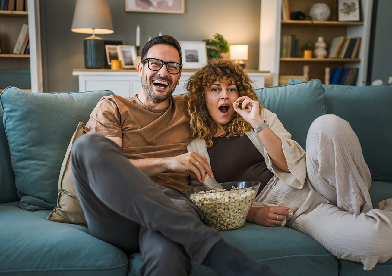 Couple man and woman caucasian husband and wife or boyfriend and girlfriend sit on the sofa bed at home watch tv movie series with bowl of popcorn happy smile bonding love family concept copy space