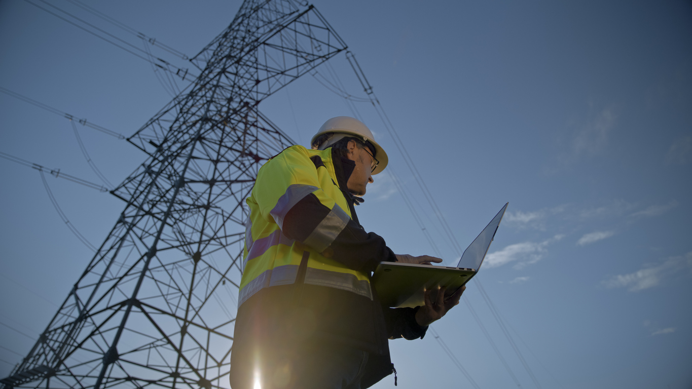 In a low angle view,a male electrical engineer gazes into the distance while holding a laptop against the backdrop of a serene blue sky. His contemplative moment amidst technology and nature