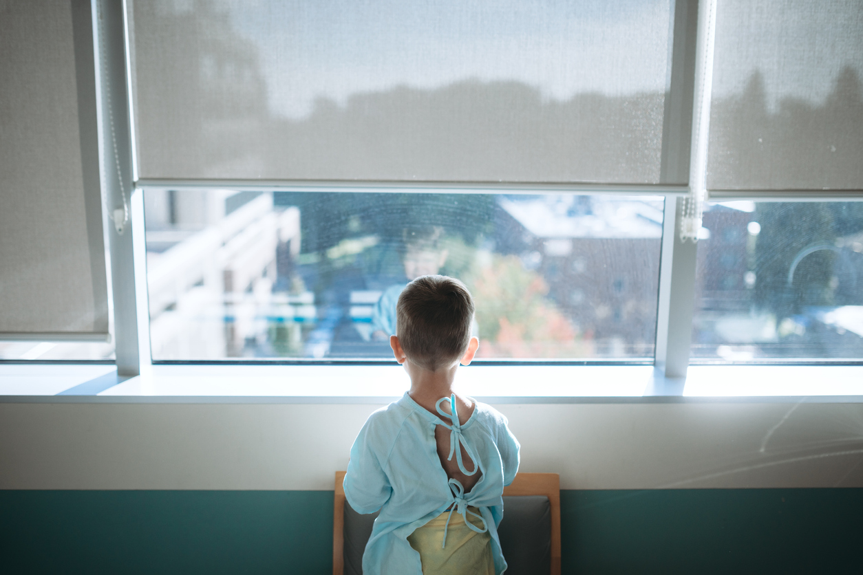 A cute little Caucasian boy waits in the childrens hospital ward to be taken back for surgery.  He looks out of the window of his room while waiting for the doctor to arrive.