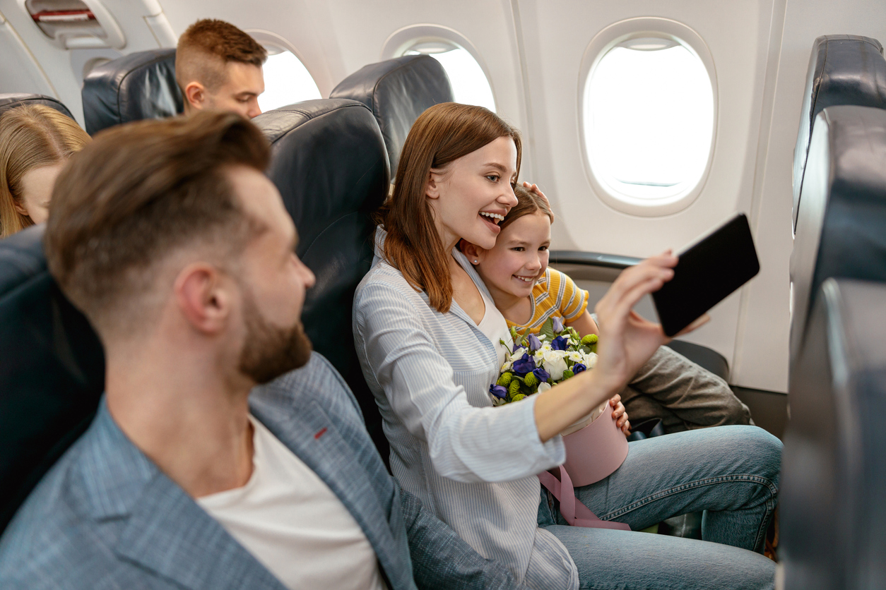 Smiling woman hugging daughter and taking picture with smartphone while sitting next to husband in passenger plane