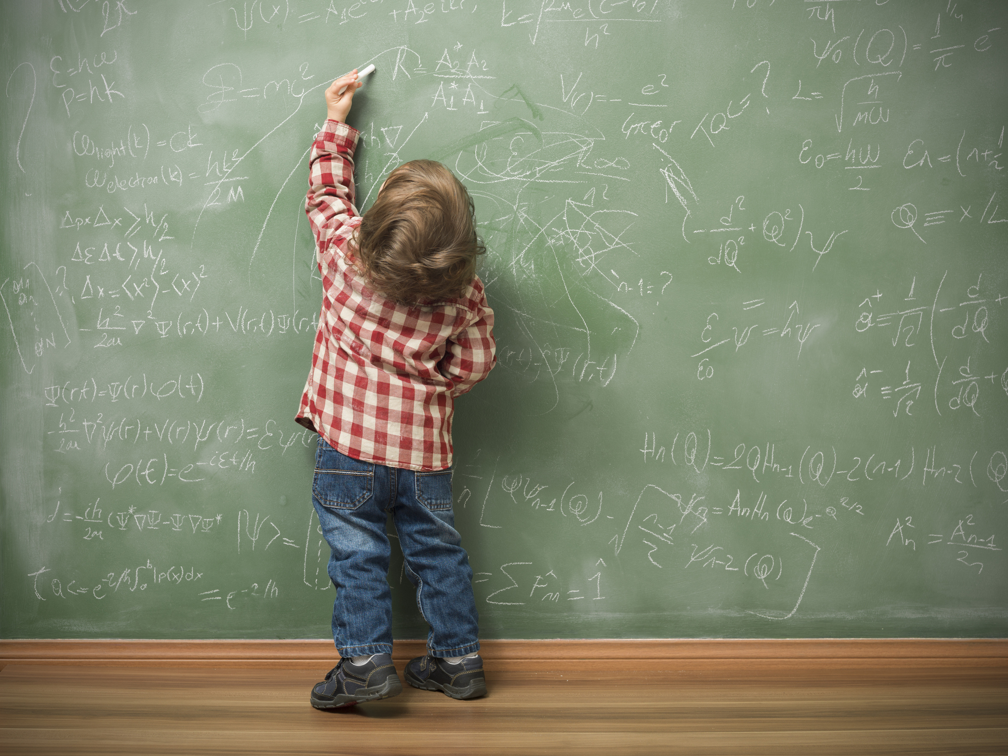 Little boy writing on green blackboard with math formulas written on.He is wearing a red plaid shirt and standing on the left side of frame.The green board is full of mathematics and physics formulas.He is seen in full length and holding chalk in left hand.The photo was shot in studio with a medium format camera Hasselblad H4D.
