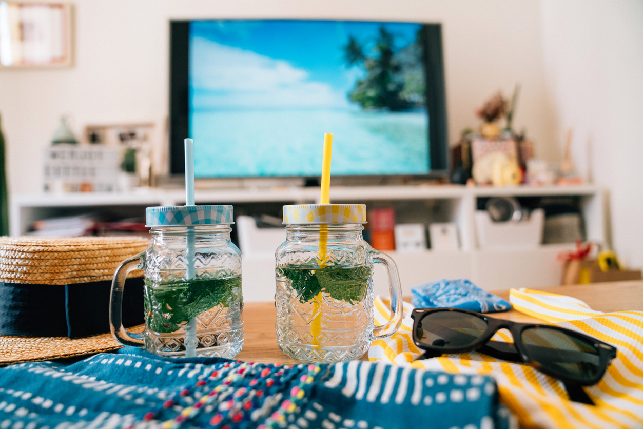 Conceptual shot of two drink glasses with a paradisiac beach on the TV screen at home. Staycation concept.