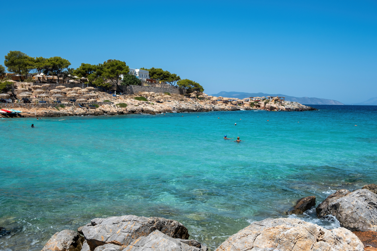 Greece Aponisos beach, destination Agistri island. Rocky beach with pine tree and umbrella, people swim in turquoise sea water, blue sky, sunny day.