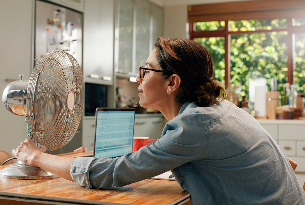 Woman, fan and laptop in home with heat wave as freelance copywriting for remote work, email or kitchen. Female person, over heating and hot weather in apartment with appliance, cool air or summer