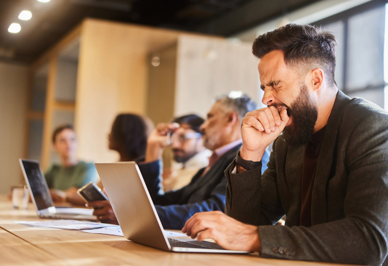Young businessman working on a laptop and yawning while sitting with coworkers at a table in an office lounge