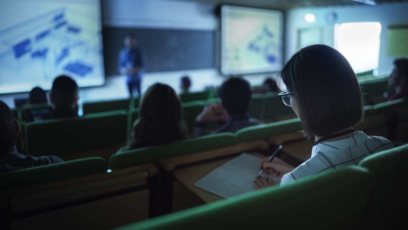 Over the Shoulder Footage of a Female Student Taking Down Notes in a Notebook, Sitting in College Auditorium on a Lecture. Young Woman with Short Hair Wearing Glasses and Studying in University