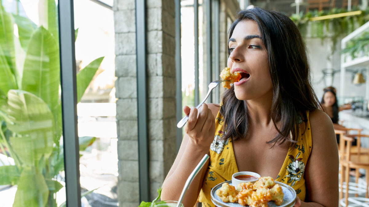 Unposed, natural shot of pretty brunette ethnic vegan model eating tasty fine vegetarian plant-based cuisine on artisanal ceramic plate including fried cauliflower and sauce with a green smoothie and reusable metal straw for mindful eating and a nutritious paleo diet.