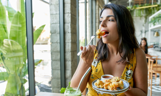 Unposed, natural shot of pretty brunette ethnic vegan model eating tasty fine vegetarian plant-based cuisine on artisanal ceramic plate including fried cauliflower and sauce with a green smoothie and reusable metal straw for mindful eating and a nutritious paleo diet.
